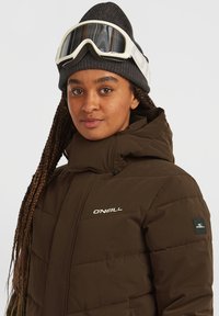 Bruine gewatteerde jas met hoge kraag, logo op de borst en een patch op de mouw. Gepaard met een grijze beanie en witte skibril.