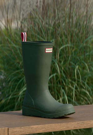 Stivale da pioggia Green Hunter con linguetta di tiraggio rossa, bianca e nera, posato su una superficie di legno con sfondo di erba alta.