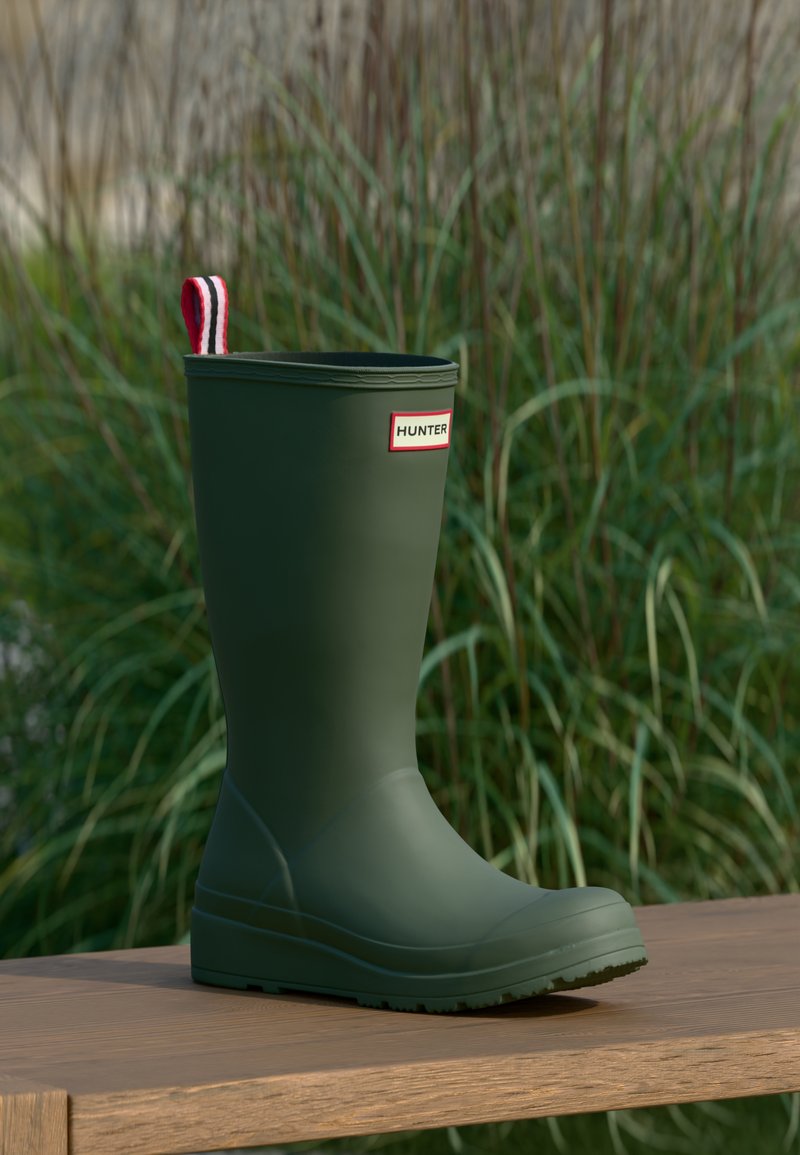 Πράσινο ανθεκτικό μπότες βροχής Hunter με έλξη σε κόκκινο, λευκό και μαύρο χρώμα, στέκεται σε ξύλινη επιφάνεια με φόντο ψηλό γρασίδι.