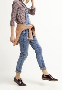 Salopette en denim délavé, portée sur une chemise à motif floral. Un pull en tricot orange noué à la taille. Chaussures brillantes bordeaux.