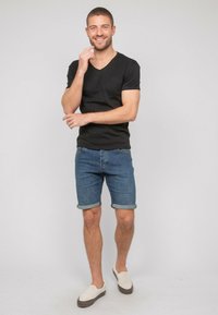 T-shirt noir à col en V, shorts en denim retroussés au niveau de l'ourlet, et chaussures légères à enfiler avec une semelle sombre, sur fond neutre.