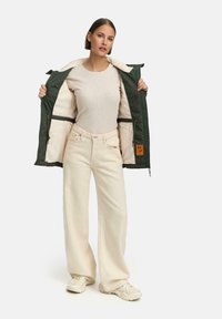 Green padded jacket with cream lining, worn open over a beige ribbed top and cream wide-leg pants. Cream sneakers complete the outfit.