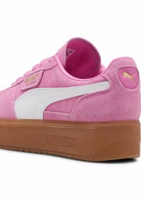 Růžová semišová teniska Puma s bílým pruhem, gumovou podrážkou, zlatým logem Puma na patě a boku, z pohledu zezadu a z boku.