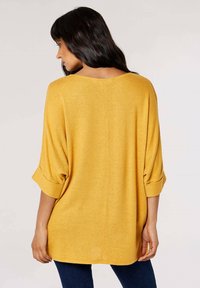 Haut à manches longues, oversize en tricot doux et texturé couleur moutarde, avec des manches retroussées et un col large. Présente un ourlet arrondi pour un ajustement décontracté.