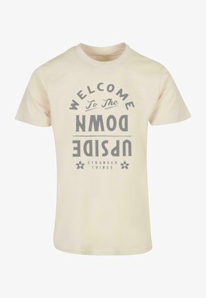 Cremfarbendes Baumwoll-T-Shirt mit einer gebogenen Grafik in grauem Text, die "WILLKOMMEN IM UPSIDE DOWN" und "STRANGER THINGS" zeigt.