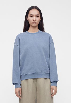 Femme aux longs cheveux noirs portant un sweat-shirt bleu ample et un pantalon beige à jambes larges sur un fond blanc uni.