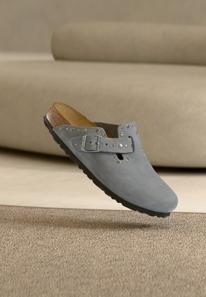 Γκρι σουέτ παπούτσι τύπου slip-on με πόρπη και μεταλλικά τρουκς, φετινή σόλα και μαύρη εξωτερική σόλα, εμφανίζεται πάνω σε μπεζ χαλί και καναπέ.
