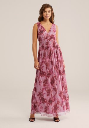 Femeie purtând o rochie lungă fără mâneci, înflorată, în nuanțe de roz și roșu, cu decolteu în V și fustă plisată, încălțată cu pantofi negri cu vârf deschis, stând pe un fundal simplu.