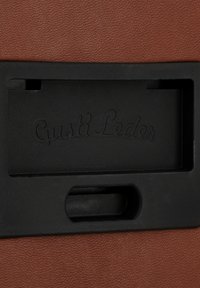 Brūna ādas izstrādājums ar teksturētu virsmu. Tam ir melna plastmasas daļa ar iegrimu un iespiestu "Gusti Leder" logotipu.