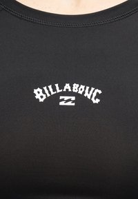 Μαύρο πουκάμισο με το λευκό όνομα και λογότυπο της μάρκας Billabong στο κέντρο του στήθους, ορατή ραφή στην περιοχή του λαιμού, φοριέται από ένα άτομο.