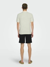 Top corto a maniche corte di colore beige chiaro con un design aderente, abbinato a pantaloni shorts neri. La texture appare liscia, con un piccolo tag laterale visibile.
