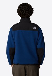 Niebieska polarowa kurtka z czarnymi akcentami, teksturowany materiał, wysoki kołnierz i logo na górnej części pleców. Posiada boczne kieszenie i luźny krój.