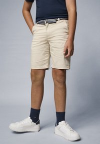 Short beige en coton avec une coupe classique, doté de poches latérales et d'une ceinture rayée noire et blanche. Associé à des baskets blanches et des chaussettes bleu marine.