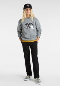Grå sweatshirt med "VANS"-grafik, gul midja, svarta byxor och svarta och vita rutiga slip-on skor. En svart keps fulländar looken.