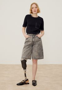 Jeune femme aux cheveux blonds courts, portant un T-shirt noir, un short en jean gris et une jambe gauche prothétique, debout avec les mains dans les poches.
