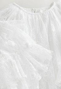 Robe en dentelle blanche avec des manches volantes, un col rond et une fermeture éclair dans le dos. La texture est délicate avec des motifs floraux.