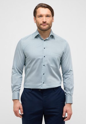 Langärmliges hellblaues Hemd mit geometrischem Muster, Knopfdown-Kragen und dunklen Knöpfen, kombiniert mit dunklen Hosen.