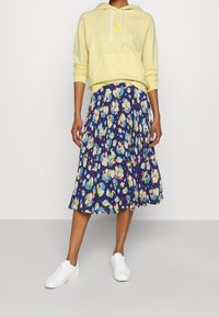 Gul hoodie med framficka och vita dragsnören, kombinerad med en blå blommig midi-kjol med veck och vita sneakers.