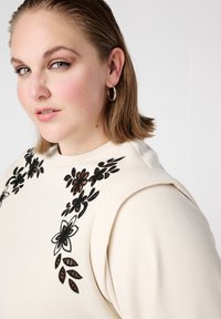 Roomkleurige sweatshirt met bloemrijke borduurwerk in zwart en bruin. Heeft een ronde halslijn en gestructureerde schouders.