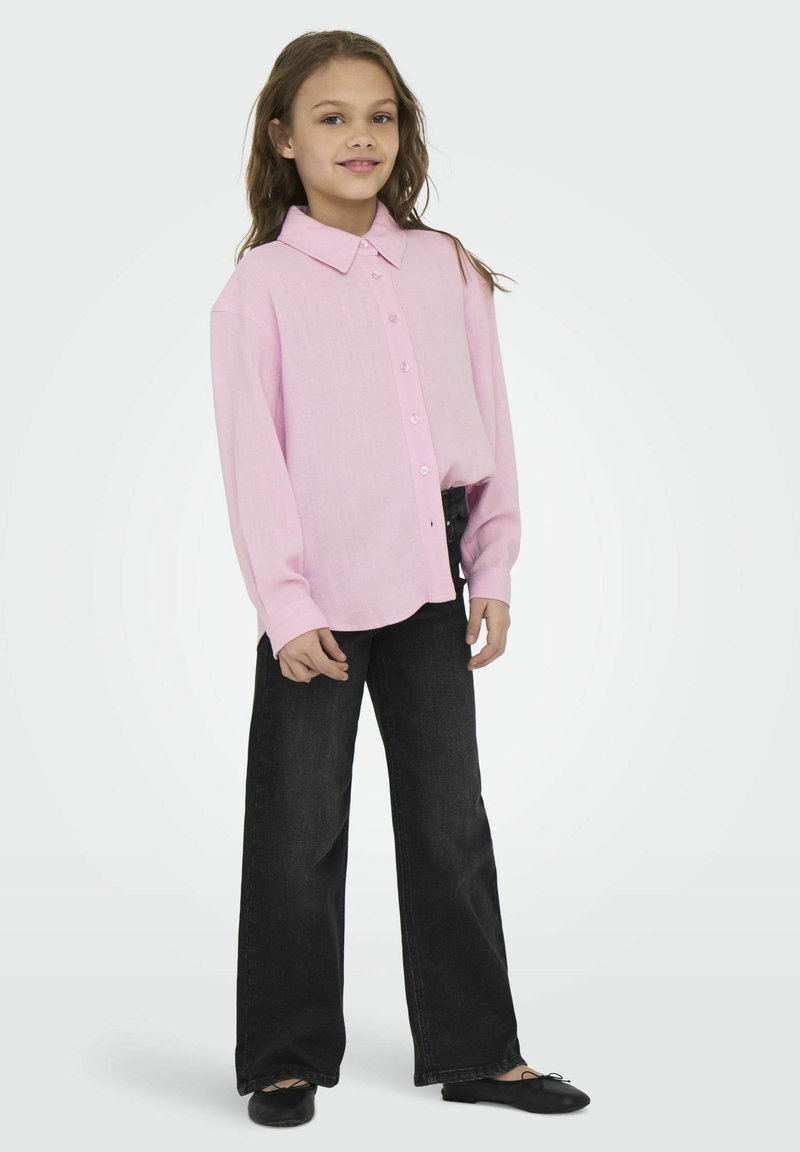 Chemise boutonnée rose pâle à manches longues, associée à un jean noir à jambes larges. La chemise présente un col classique et une texture subtile.