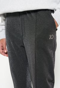 Zwarte, glanzende trainingbroek met een subtiele glittersffect. Kenmerken zijn onder andere zijzakken en een geborduurd '10' op de voorkant. Gladde textuur.
