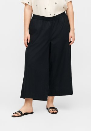 Γυναίκα που φορά χαλαρά μαύρα φαρδιά παντελόνια μέχρι τον αστράγαλο, μπεζ κοντομάνικο πουκάμισο με σχέδια φοίνικες και μαύρα σανδάλια χωρίς λουράκια με χρυσή λεπτομέρεια.