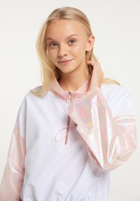 Hvid let pullover med pastelfarvede lyserøde iriserende ærmer og krave. Har en lynlås foran og snoredetalje ved halsen.