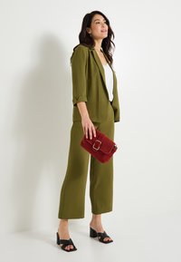Costume vert olive avec une coupe décontractée, associé à un haut blanc, des sandales à talons carrés noires et une pochette en daim rouge avec une boucle en or.