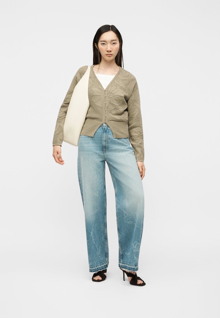 Mujer con cárdigan beige con estampado, vaqueros de pierna ancha azul claro, tacones negros y un gran bolso blanco de hombro sobre fondo blanco liso.
