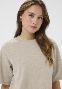 Bežs īsām piedurknēm T-krekls mīkstā audumā ar apaļu kakla izgriezumu. Modelim ir gaiši brūni mati un viņš valkā zelta riņķveida auskarus.