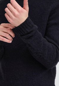 Pulover închis la culoare, cu o suprafață texturată și manșete cu model. Prim-plan cu o mână care ajustează mânecă. Materialul pare moale și călduros.