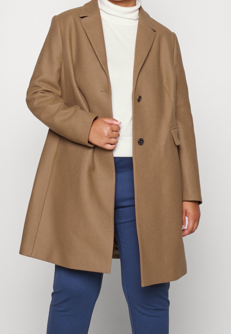 Brown wool-blend coat with notched lapels, double-breasted buttons, and side pockets, paired with a light turtleneck and blue trousers.