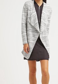 Grå och vit mönstrad cardigan med öppen front, långa ärmar och mjuk textur, lager på lager över en svart klänning.
