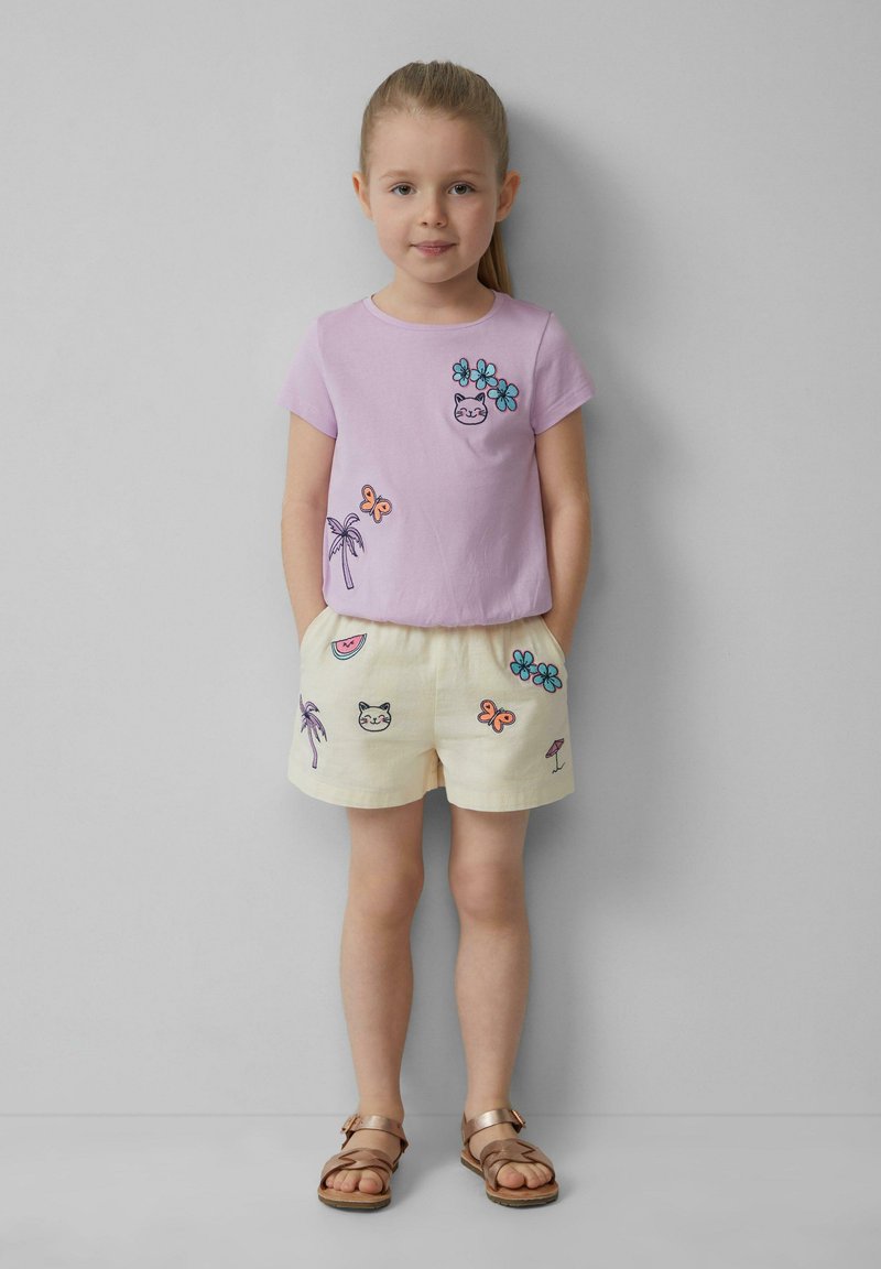 Lila T-shirt met korte mouwen, versierd met bloemen- en kattenborduurwerk, gecombineerd met crèmekleurige shorts met fruit- en vlinderontwerpen. Bruine sandalen.