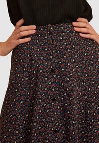 Gonna a forma A floreale con sfondo scuro, decorata con piccoli fiori rossi, blu e gialli, e bottoni in ottone lungo la parte anteriore. Presenta una vita testurizzata.