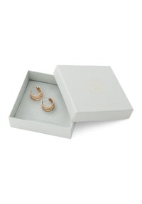 Goldfarbene Creolen in einer hellgrauen, strukturierten Schachtel mit dezentem Logo. Sie haben ein wellenförmiges Design und einen sicheren Steckverschluss.