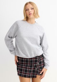 Sudadera gris claro con mangas raglán, combinada con una falda mini de cuadros negros y rojos que presenta botones laterales y un dobladillo estructurado.