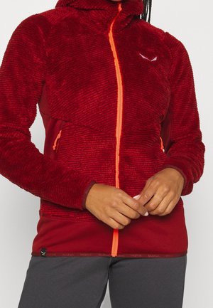 Personne portant une veste en polaire rouge texturée avec une fermeture éclair orange et des poches latérales, mains jointes à la taille, pantalon foncé visible.