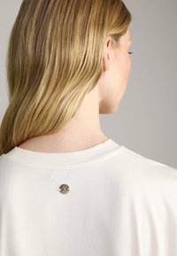 Weißes Baumwoll-T-Shirt mit runder silberner Knopfdetails am Nacken. Glatte Textur und schlichtes Design.