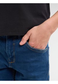 Modré džíny z denimu se žlutým prošíváním, s přední kapsou. Tkanina vypadá hladce a má klasický střih.