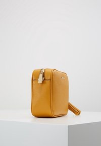 Gul læder kosmetikpung med struktureret overflade, lynlåslukning og en sidekvast. Firkantet form, kompakt design.