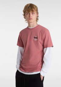 Camiseta de algodón rosa con mangas cortas, que presenta un logo negro de Vans y una camisa blanca de manga larga superpuesta. Pantalones negros lisos.