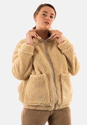 Veste en polaire marron clair avec un col montant, fermeture éclair à l'avant et deux grandes poches. Présente une surface douce et texturée ainsi qu'une coupe décontractée.
