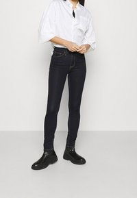 Tmavé denim skinny džíny s vysokým pasem, zlatým prošíváním a hladkou texturou, spárované s bílou košilí na knoflíky a černými botami.