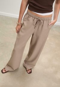 Beige wijde broek met een elastische tailleband en trekkoord, voorzien van zijzakken. Gepaard met bruine teenslippers en gelaagde armbanden.
