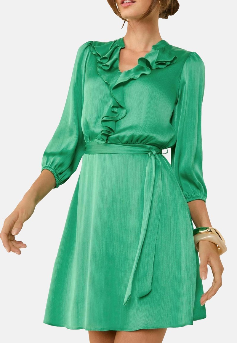 Vestido de satén verde con escote volante y mangas largas. La falda acampanada presenta un cinturón atado, creando una silueta suave y fluida.