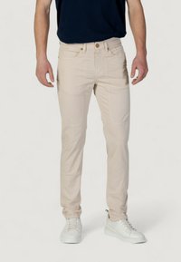 Pantalones beige de corte slim con bolsillos frontales, cierre de botón y detalles de costura sutiles. Combinados con zapatillas blancas y una camiseta azul marino.