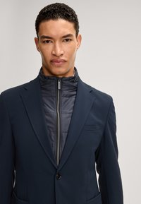 Marineblazer met gestructureerde revers over een donkere pufferjas. Beschikt over een ritssluiting aan de voorkant, horizontale gestikte textuur en een voorzak.