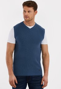 Navyblauwe gebreide mouwloze trui met een V-hals ontwerp, gedragen over een wit shirt met korte mouwen, gecombineerd met blauwe spijkerbroeken. Getextureerde stof.