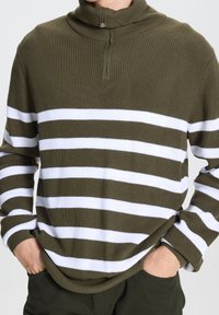 Persona con un suéter de punto a rayas verde olivo y blanco, con un cuello de media cremallera y manos en los bolsillos de unos pantalones verdes oscuros.
